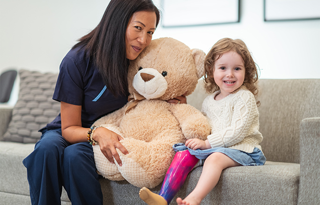 Therapist and pediatric patient with pink prosthetic leg sitting on couch holding teddy bear